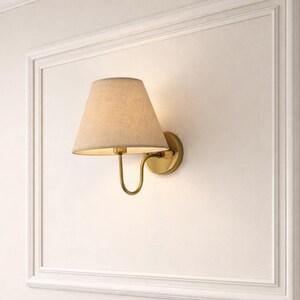 May include: A wall-mounted lamp with a beige lampshade and a brass-colored base. The lamp is set against a white wall within a white framed border, creating a classic and elegant look. The lamp's design is simple yet stylish.