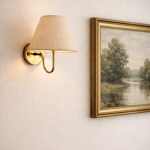 May include: A wall-mounted brass sconce with a beige lampshade. The sconce has a curved arm and a circular base. A framed landscape painting with a gold frame hangs to the right of the sconce.