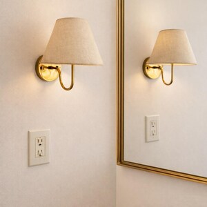 May include: A pair of wall sconces with beige fabric shades and brass-colored metal fixtures. The sconces are mounted on a white wall, with a matching electrical outlet below. A mirror with a gold frame is visible.