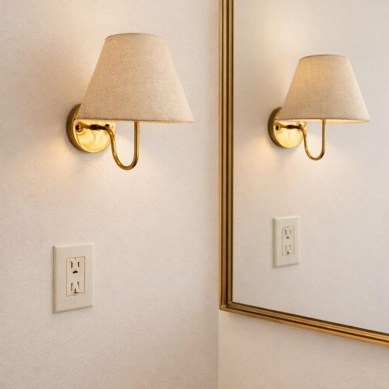 May include: A pair of wall sconces with beige fabric shades and brass-colored metal fixtures. The sconces are mounted on a white wall, with a matching electrical outlet below. A mirror with a gold frame is visible.