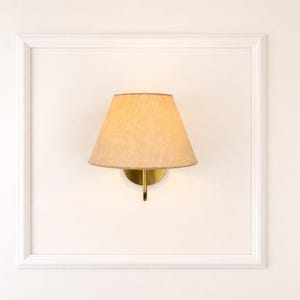 May include: A wall-mounted lamp with a beige lampshade and a brass-colored base. The lamp is set within a white, rectangular frame, creating a clean and minimalist aesthetic. The design is suitable for interior decor.