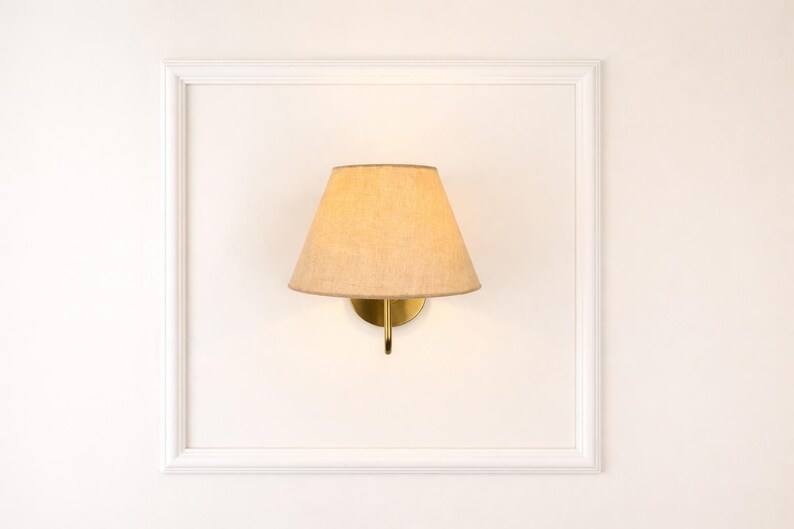 May include: A wall-mounted lamp with a beige lampshade and a brass-colored base. The lamp is set within a white, rectangular frame, creating a clean and minimalist aesthetic. The design is suitable for interior decor.