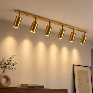 May include: A ceiling-mounted track lighting fixture featuring a long, straight bar in a brushed brass finish with six cylindrical spotlights attached. Each spotlight is oriented downward, emitting a warm, focused glow onto the wall below.