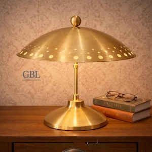 以下が含まれることがあります： ドーム型のシェードに小さな穴が等間隔に開いた真鍮色のデスクランプ。 ランプは丸いベースと細いステムを備えています。 本とメガネが置かれた木製の表面にあります。