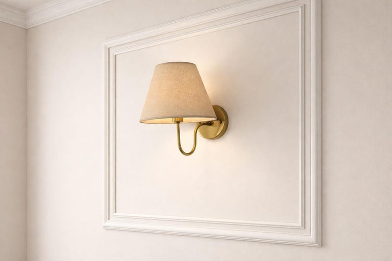 May include: A wall-mounted lamp with a beige lampshade and a brass-colored base. The lamp is set against a white wall within a white framed border, creating a classic and elegant look. The lamp's design is simple yet stylish.