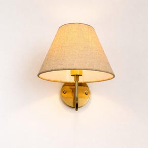 May include: A wall-mounted sconce with a beige fabric shade and a brass-colored metal base. The shade is cone-shaped, and the fixture is designed to be mounted on a wall. The light fixture is a classic design.