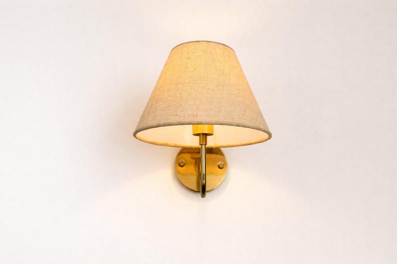 May include: A wall-mounted sconce with a beige fabric shade and a brass-colored metal base. The shade is cone-shaped, and the fixture is designed to be mounted on a wall. The light fixture is a classic design.