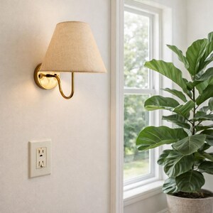 May include: A wall-mounted brass sconce with a beige linen shade. The fixture has a curved arm and a circular base. The sconce is mounted on a white wall, with a power outlet visible below. A large potted plant is in the background.