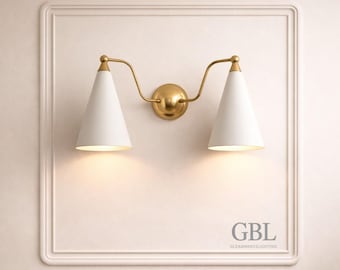 May include: A wall-mounted light fixture with two white, cone-shaped shades and a gold-colored frame. The fixture is mounted on a cream-colored wall with a decorative frame. The text "GBL GLEAMBRASSLIGHTING" is visible.