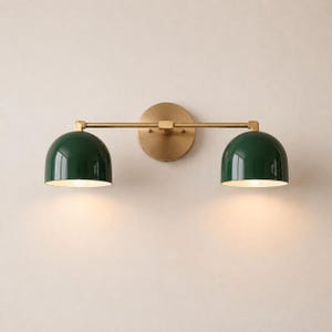 Può includere: Una lampada da parete con una barra orizzontale in ottone fissata a una piastra circolare in ottone. Due paralumi a cupola verde scuro sono posizionati alle estremità della barra, ciascuno contenente una lampadina che emette una luce calda.