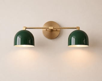 May include: A wall-mounted light fixture featuring a horizontal brass bar attached to a circular brass backplate. Two dark green dome-shaped shades are positioned at each end of the bar, directing light downward from the interior bulbs.