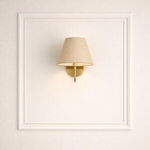 May include: A wall-mounted sconce with a beige fabric shade and a gold-colored metal base. The sconce is mounted on a white wall within a white rectangular frame, providing a classic and elegant lighting solution.