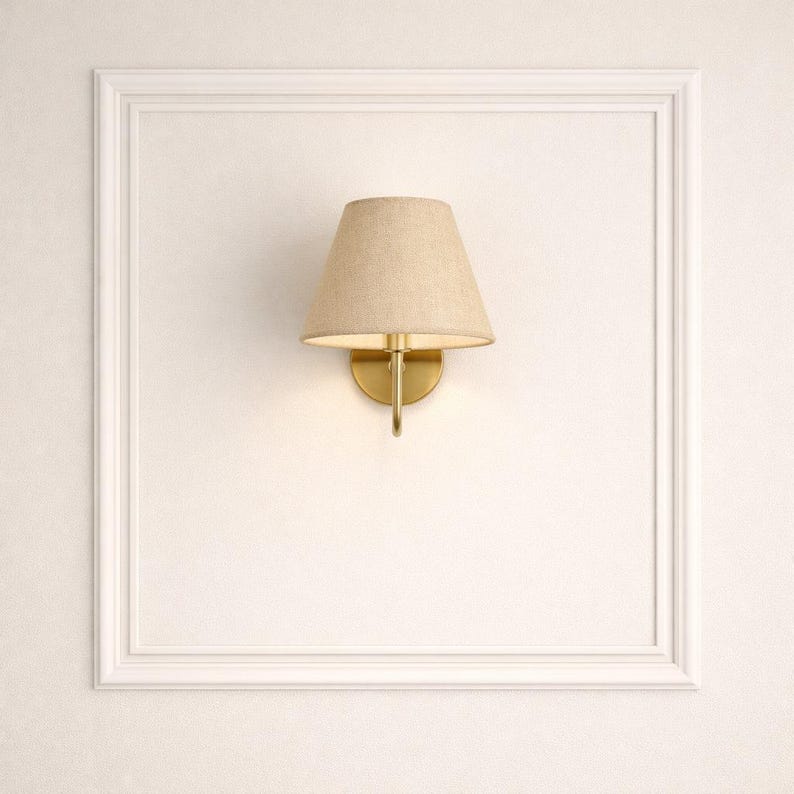 May include: A wall-mounted sconce with a beige fabric shade and a gold-colored metal base. The sconce is mounted on a white wall within a white rectangular frame, providing a classic and elegant lighting solution.