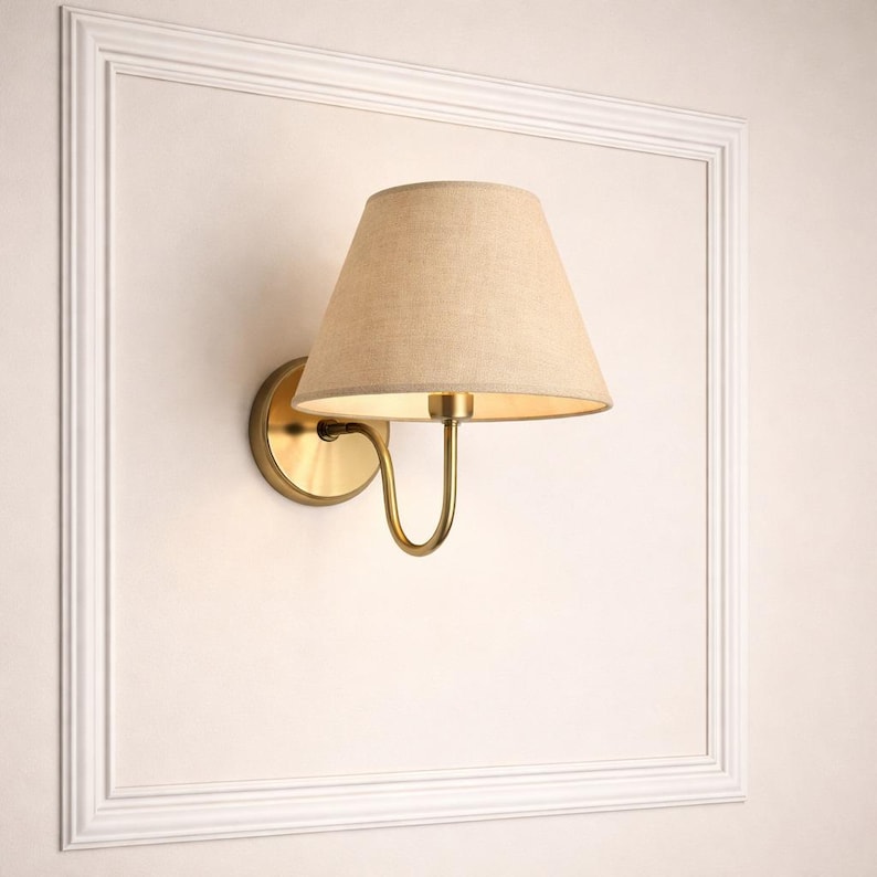 May include: A wall-mounted sconce with a beige fabric shade and a brass-colored metal base. The sconce is mounted on a white wall within a white framed border. The lamp's design is simple and elegant, suitable for various interior styles.