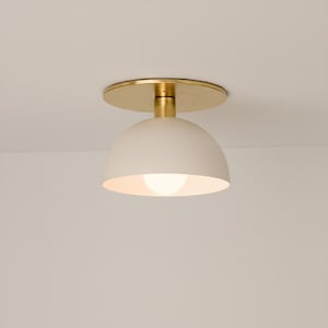 Pode incluir: Uma moderna luminária de teto com uma cúpula branca e uma base e placa de montagem douradas. A lâmpada é visível dentro da cúpula. A luminária é montada em um teto branco.