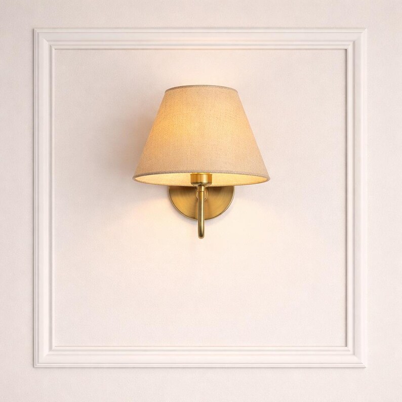 May include: A wall-mounted sconce with a beige fabric shade and a brass finish. The sconce has a classic design, with a conical shade and a curved arm. The light fixture is mounted on a white wall with a decorative frame.