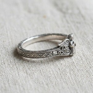 May include: A silver ring featuring an intricate band with a repeating cross-hatch pattern and a small embedded stone. The ring is topped with a sculpted figure of an angel with detailed wings and a small cross motif integrated into the metalwork.