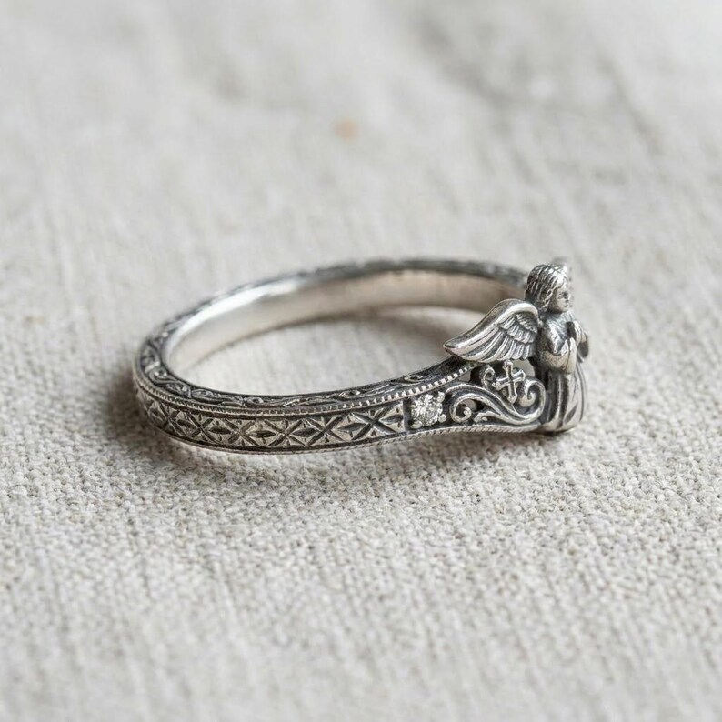 May include: A silver ring featuring an intricate band with a repeating cross-hatch pattern and a small embedded stone. The ring is topped with a sculpted figure of an angel with detailed wings and a small cross motif integrated into the metalwork.