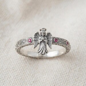 Puede incluir: Un anillo de plata con una figura central de &aacute;ngel con las alas extendidas y las manos juntas en oraci&oacute;n. La banda est&aacute; decorada con patrones florales grabados, una peque&ntilde;a cruz y tres piedras preciosas rojas redondas incrustadas.