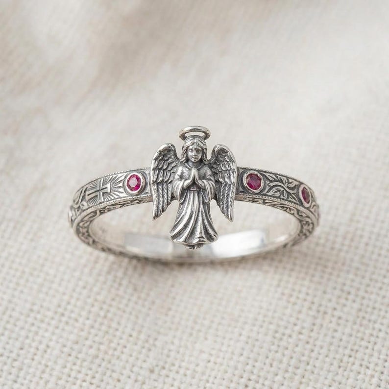 Puede incluir: Un anillo de plata con una figura central de &aacute;ngel con las alas extendidas y las manos juntas en oraci&oacute;n. La banda est&aacute; decorada con patrones florales grabados, una peque&ntilde;a cruz y tres piedras preciosas rojas redondas incrustadas.