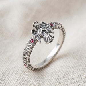 Puede incluir: Un anillo de plata que presenta una figura de &aacute;ngel tridimensional con alas y halo. La banda est&aacute; decorada con patrones florales grabados, un peque&ntilde;o s&iacute;mbolo de cruz y varias piedras preciosas rojas, redondas y peque&ntilde;as incrustadas.