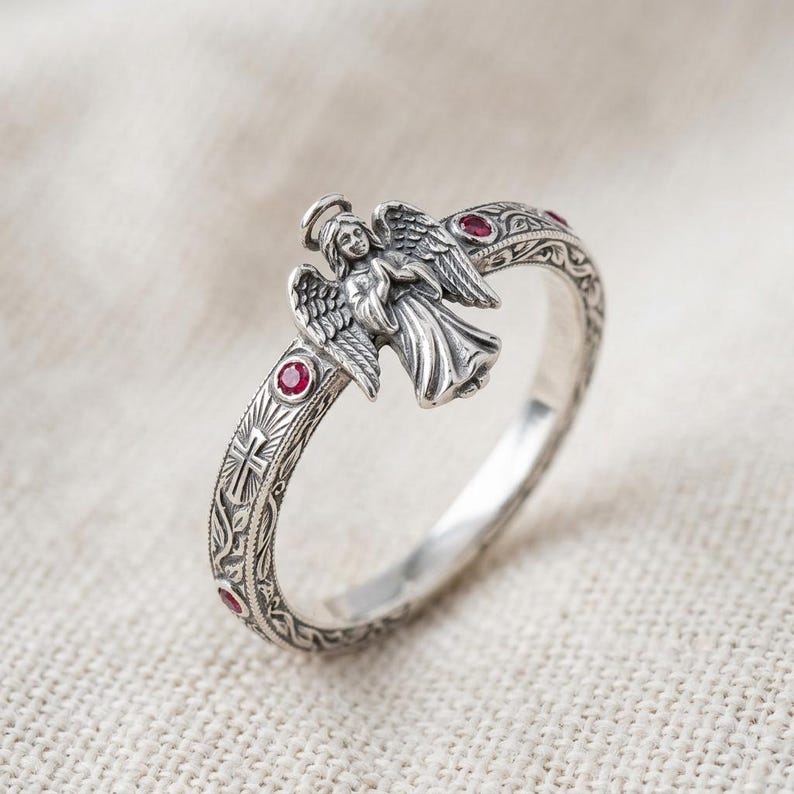 Puede incluir: Un anillo de plata que presenta una figura de &aacute;ngel tridimensional con alas y halo. La banda est&aacute; decorada con patrones florales grabados, un peque&ntilde;o s&iacute;mbolo de cruz y varias piedras preciosas rojas, redondas y peque&ntilde;as incrustadas.