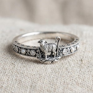 May include: A silver ring featuring a sculpted lamb standing next to a shepherd's crook. The band is decorated with an intricate engraved pattern, small round white pearls, and clear sparkling stones set along the outer circumference.