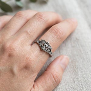 May include: A silver-toned metal ring featuring a central angel figure with outstretched wings. The band is decorated with intricate scrollwork and small cross motifs, showcasing an oxidized finish that highlights the detailed relief design.