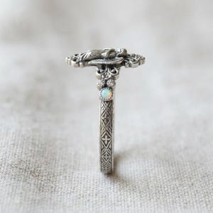 May include: A silver ring featuring an intricate angel figure at the top, accented with a small round iridescent opal stone and tiny clear stones. The band displays a detailed cross-hatch pattern along the sides, finished in an oxidized silver.