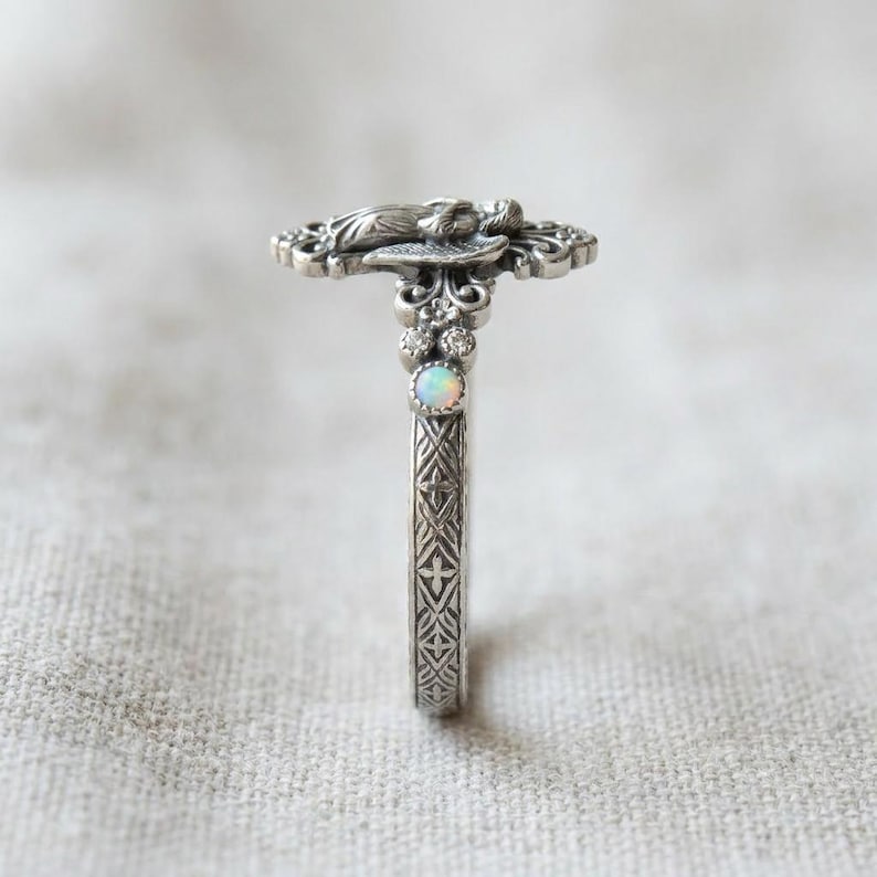 May include: A silver ring featuring an intricate angel figure at the top, accented with a small round iridescent opal stone and tiny clear stones. The band displays a detailed cross-hatch pattern along the sides, finished in an oxidized silver.