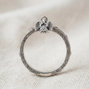 Puede incluir: Un anillo de plata que presenta una figura de &aacute;ngel esculpida con las manos juntas en la parte superior. La banda est&aacute; decorada con un intrincado patr&oacute;n de enredadera grabado y peque&ntilde;as piedras rojas circulares incrustadas a intervalos.