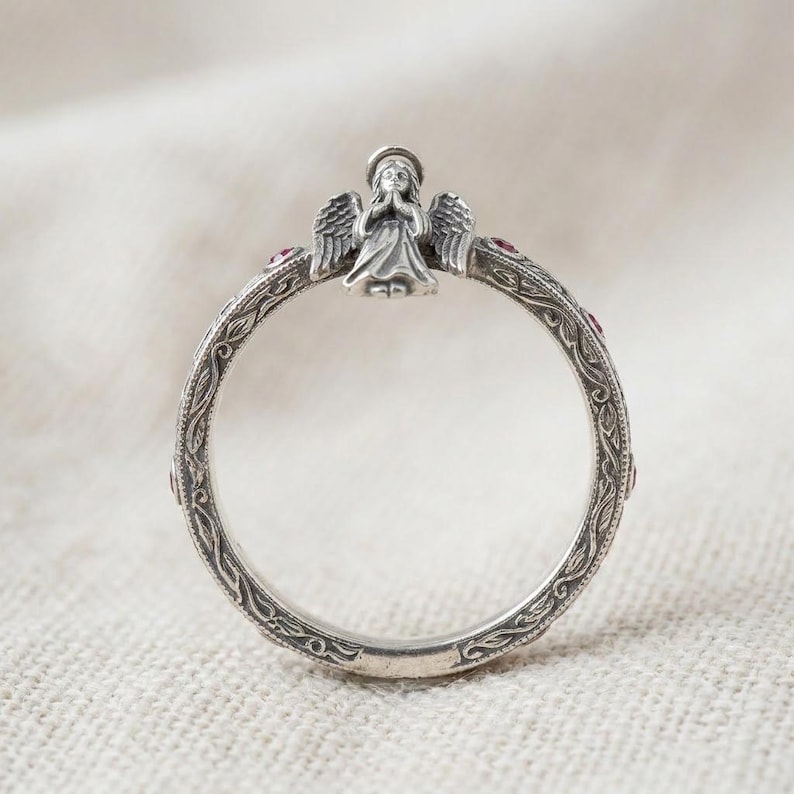 Puede incluir: Un anillo de plata que presenta una figura de &aacute;ngel esculpida con las manos juntas en la parte superior. La banda est&aacute; decorada con un intrincado patr&oacute;n de enredadera grabado y peque&ntilde;as piedras rojas circulares incrustadas a intervalos.
