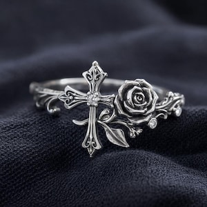 May include: A silver ring featuring a cross with a central gemstone and a rose design. The ring has intricate detailing and is set against a dark background, showcasing its gothic and floral elements.