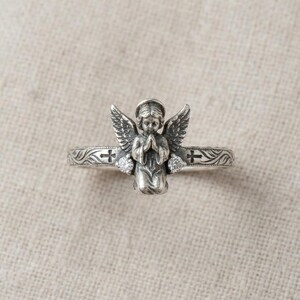 May include: A silver-toned ring featuring a central three-dimensional figure of a kneeling angel with wings spread. The band is decorated with engraved leaf patterns, two small crosses, and two clear round stones set on either side of the angel.