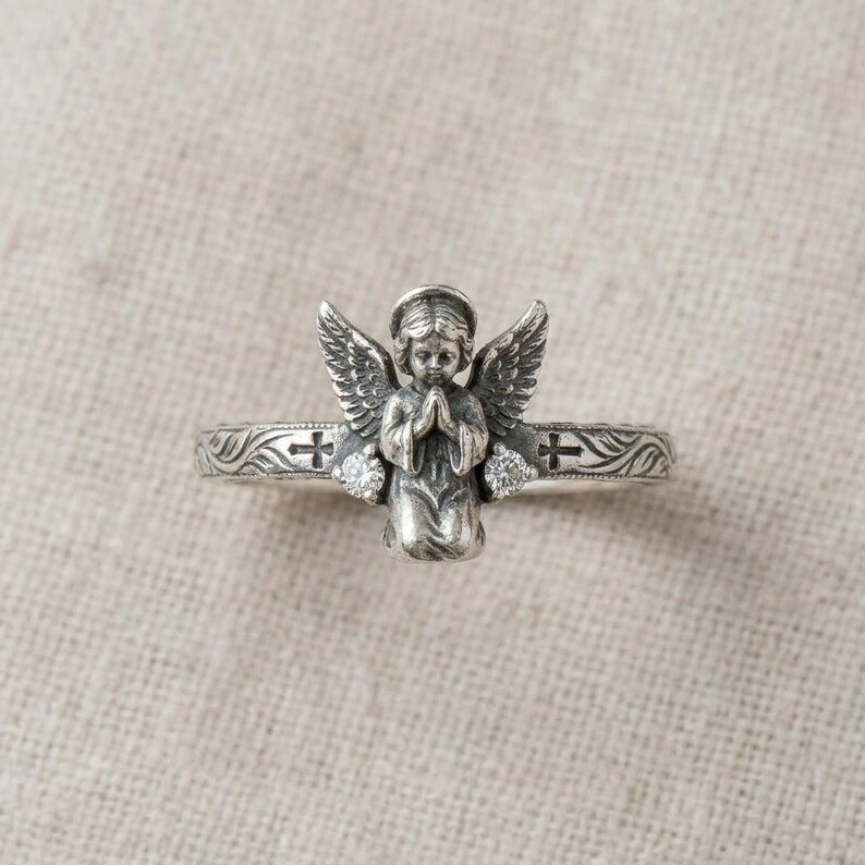 May include: A silver-toned ring featuring a central three-dimensional figure of a kneeling angel with wings spread. The band is decorated with engraved leaf patterns, two small crosses, and two clear round stones set on either side of the angel.