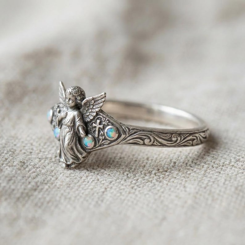 May include: A silver ring featuring a sculpted angel figure with wings, a small cross emblem, and three round iridescent opal stones. The band is decorated with intricate scrollwork engravings that extend along the sides of the metal ring.