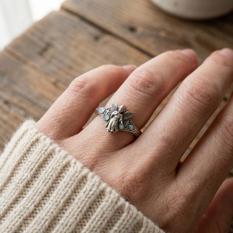 May include: A silver ring featuring a central angel figure with outstretched wings, flanked by two small crosses and four round, light blue iridescent stones set into the band. The metal has an oxidized finish to highlight the intricate details.