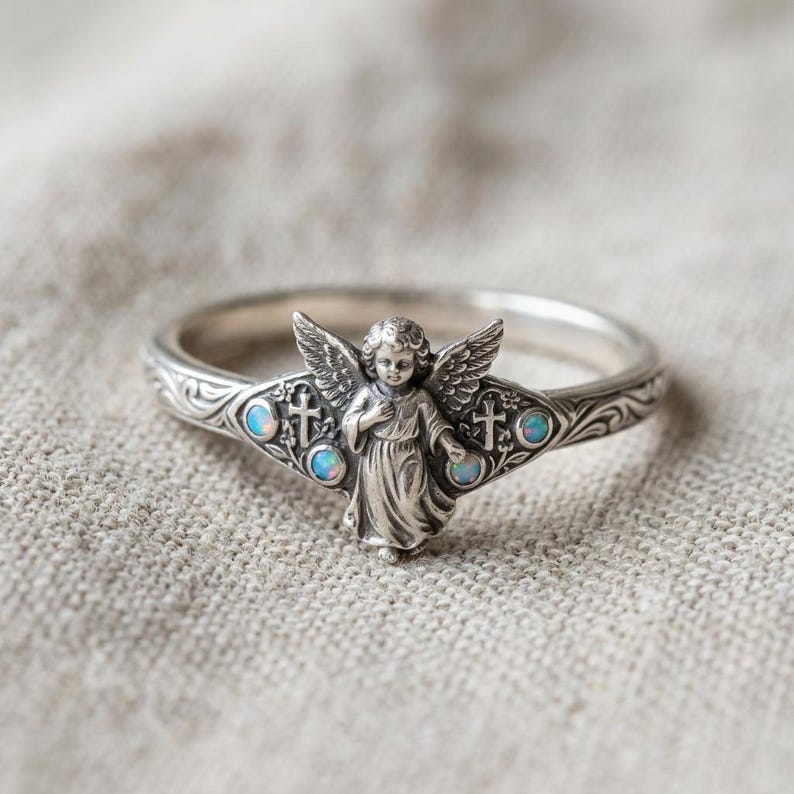 May include: A silver ring featuring a central cherub figure with wings, flanked by two small crosses and four round iridescent opal stones. The band is decorated with intricate scrollwork engravings that extend around the entire circumference.
