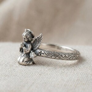 May include: A silver ring featuring a sculpted praying angel with wings, a small clear round gemstone, and a band decorated with an engraved cross and leaf patterns. The metal has an oxidized finish that highlights the intricate details.