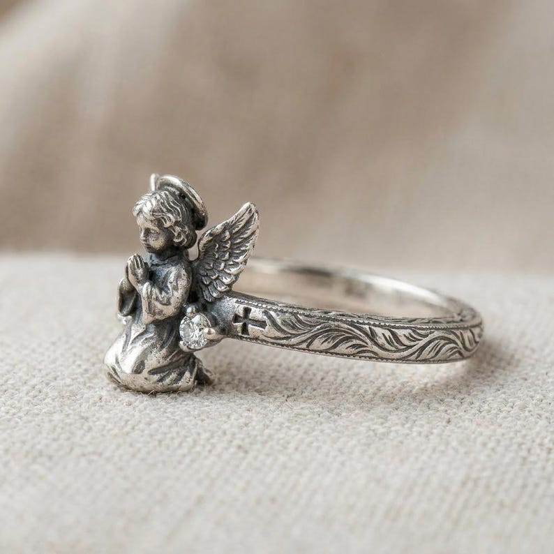 May include: A silver ring featuring a sculpted praying angel with wings, a small clear round gemstone, and a band decorated with an engraved cross and leaf patterns. The metal has an oxidized finish that highlights the intricate details.