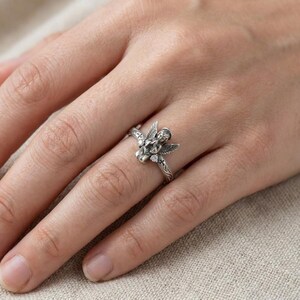 May include: A silver-toned ring featuring a sculpted angel with folded hands and wings. The band is detailed with small cross engravings and a clear, round-cut stone set on each side of the angel figure, creating a detailed metallic accessory.