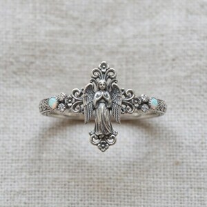 May include: A silver ring featuring a central angel figure with wings, surrounded by intricate scrollwork and floral details. The band is accented with two small, round, iridescent opals and four tiny clear stones set symmetrically on each side.