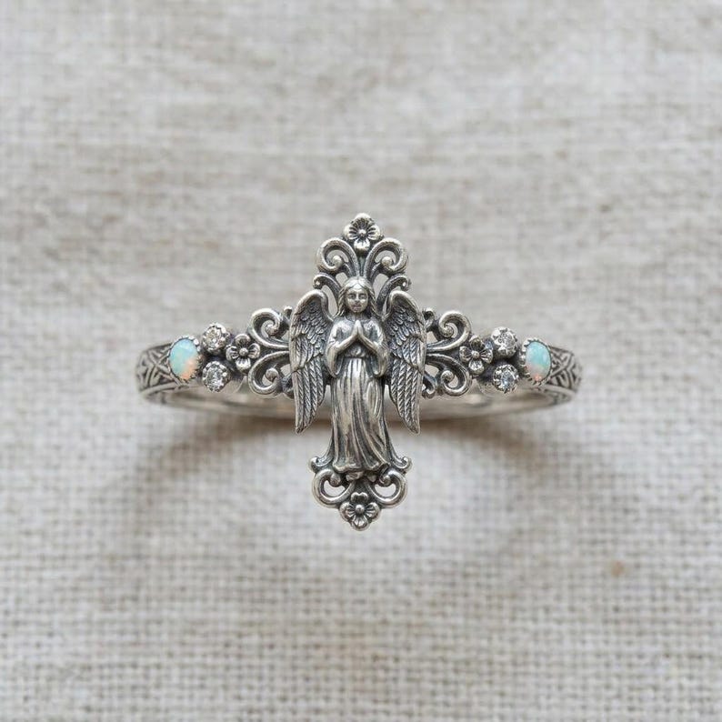 May include: A silver ring featuring a central angel figure with wings, surrounded by intricate scrollwork and floral details. The band is accented with two small, round, iridescent opals and four tiny clear stones set symmetrically on each side.