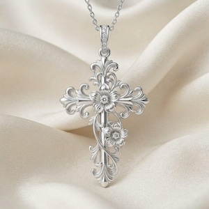 Peut inclure: Un pendentif croix en argent orné de volutes complexes et de deux motifs floraux, chacun centré par une petite pierre étincelante. La croix est suspendue à une fine chaîne en argent, présentant une finition métallique polie et des détails en filigrane.