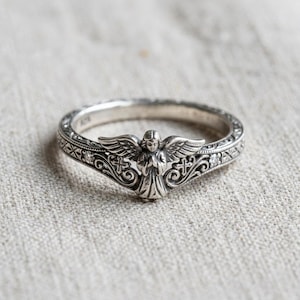 May include: A silver ring featuring a central sculpted angel figure with outstretched wings, flanked by small cross motifs and intricate scrollwork patterns. The band includes two small embedded clear stones on either side of the angel.