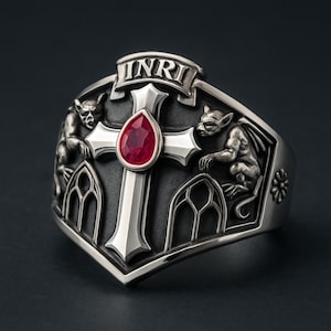 May include: Silver ring with a cross and a red gemstone, gargoyle figures, and gothic window designs. The ring has the inscription "INRI". The ring is a dark silver colour.