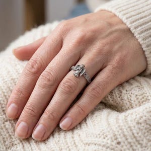 Puede incluir: Un anillo de plata que presenta una figura de &aacute;ngel detallada con alas extendidas en el centro. La banda tiene un patr&oacute;n intrincado y texturizado en los lados y est&aacute; acentuada con peque&ntilde;as piedras de color rojo oscuro a cada lado.
