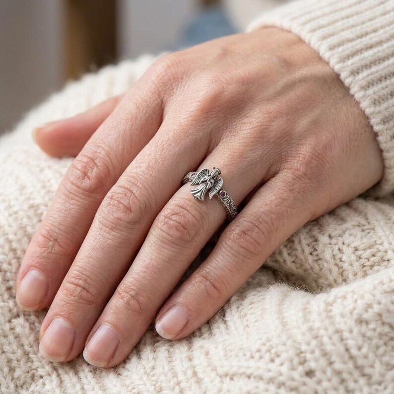 Puede incluir: Un anillo de plata que presenta una figura de &aacute;ngel detallada con alas extendidas en el centro. La banda tiene un patr&oacute;n intrincado y texturizado en los lados y est&aacute; acentuada con peque&ntilde;as piedras de color rojo oscuro a cada lado.