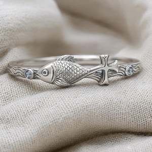 May include: A silver ring featuring a detailed fish motif with a cross-shaped tail. The band is engraved with a wavy pattern and accented by two small, round, light blue gemstones set on either side of the central fish design.
