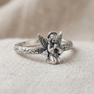 May include: A silver ring featuring a three-dimensional kneeling angel with folded hands and wings. The band is decorated with engraved leaf patterns and small cross symbols, accented by two small clear round stones at the angel's base.
